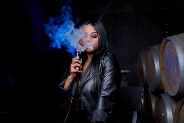 Woman in leather jacket exhaling vapor cloud from vape device in dark setting with wine barrels in background